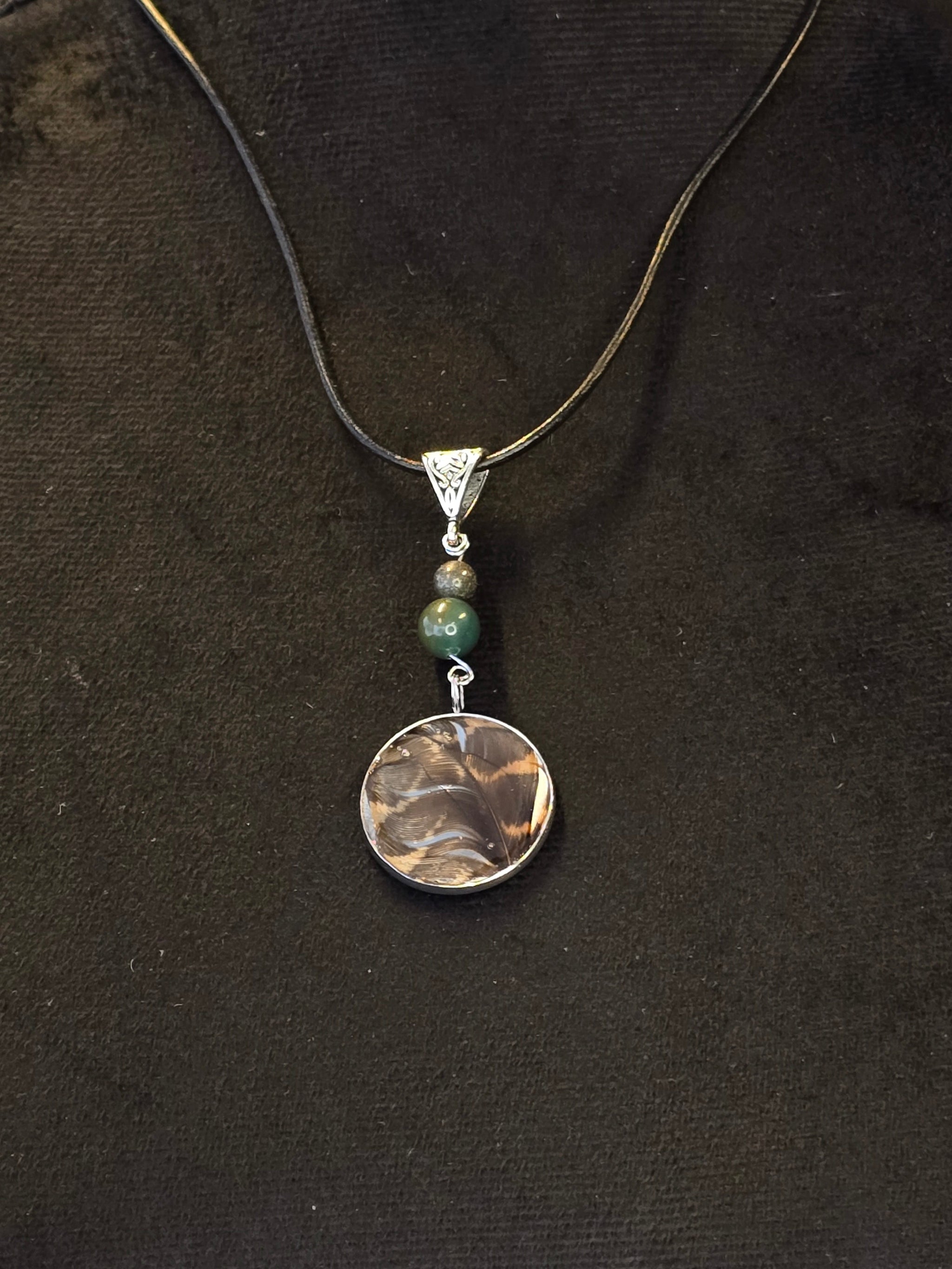 Metsän Kajo niminen amuletti, jossa on hartsi amuletin sisällä lehtokurpan sulka, ja sammalakaatti ja pyriittihelmet.