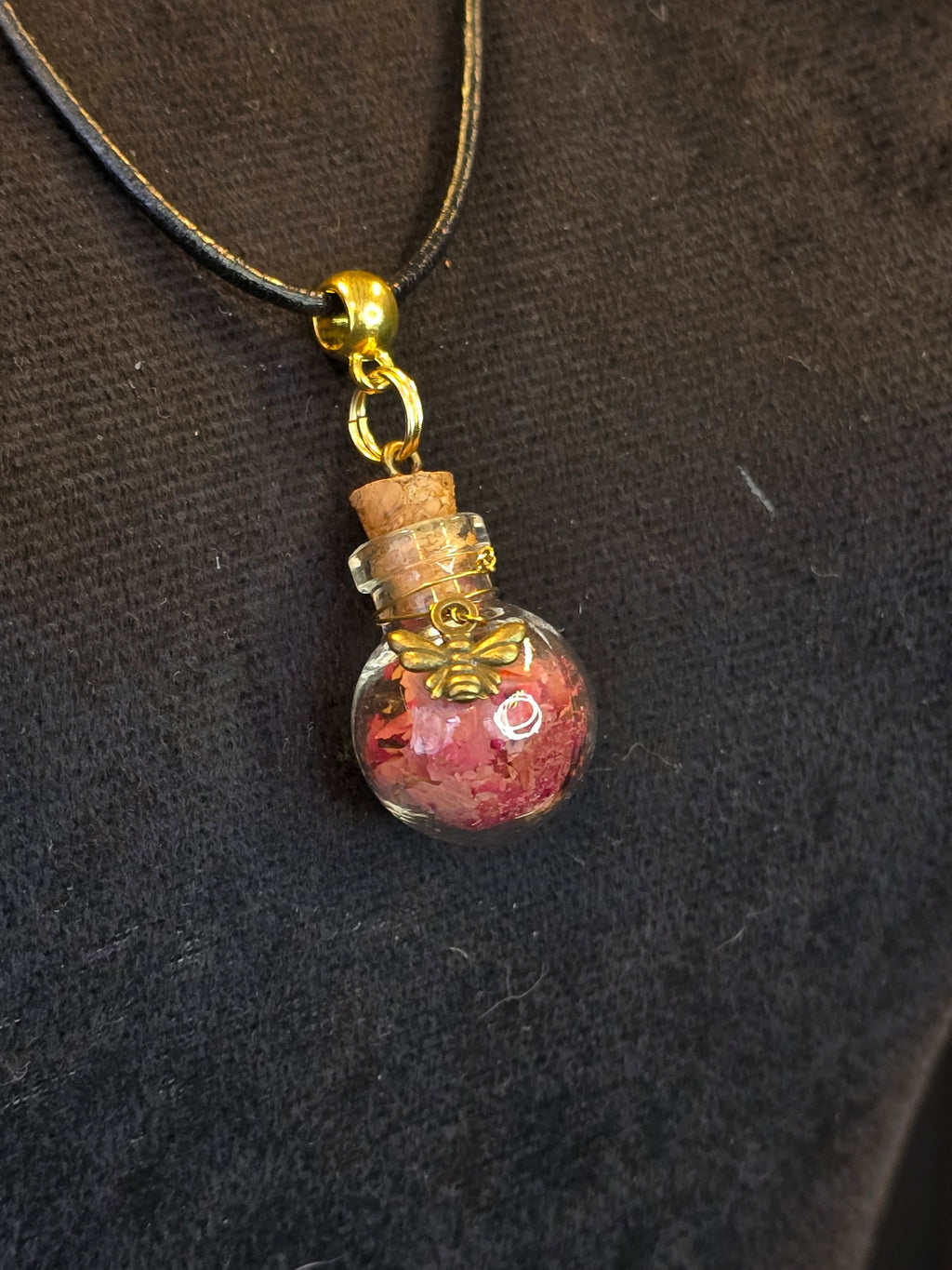 Kaulakoru, jossa lasipulloriipuksen sisällä ruusunterälehtiä ja rodoniittikivi, somisteena pieni yöperhonen riipus. 