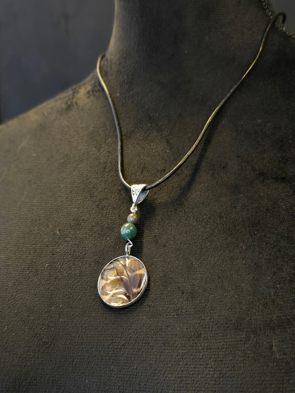 Metsän Kajo niminen amuletti, jossa on hartsi amuletin sisällä lehtokurpan sulka, ja sammalakaatti ja pyriittihelmet.