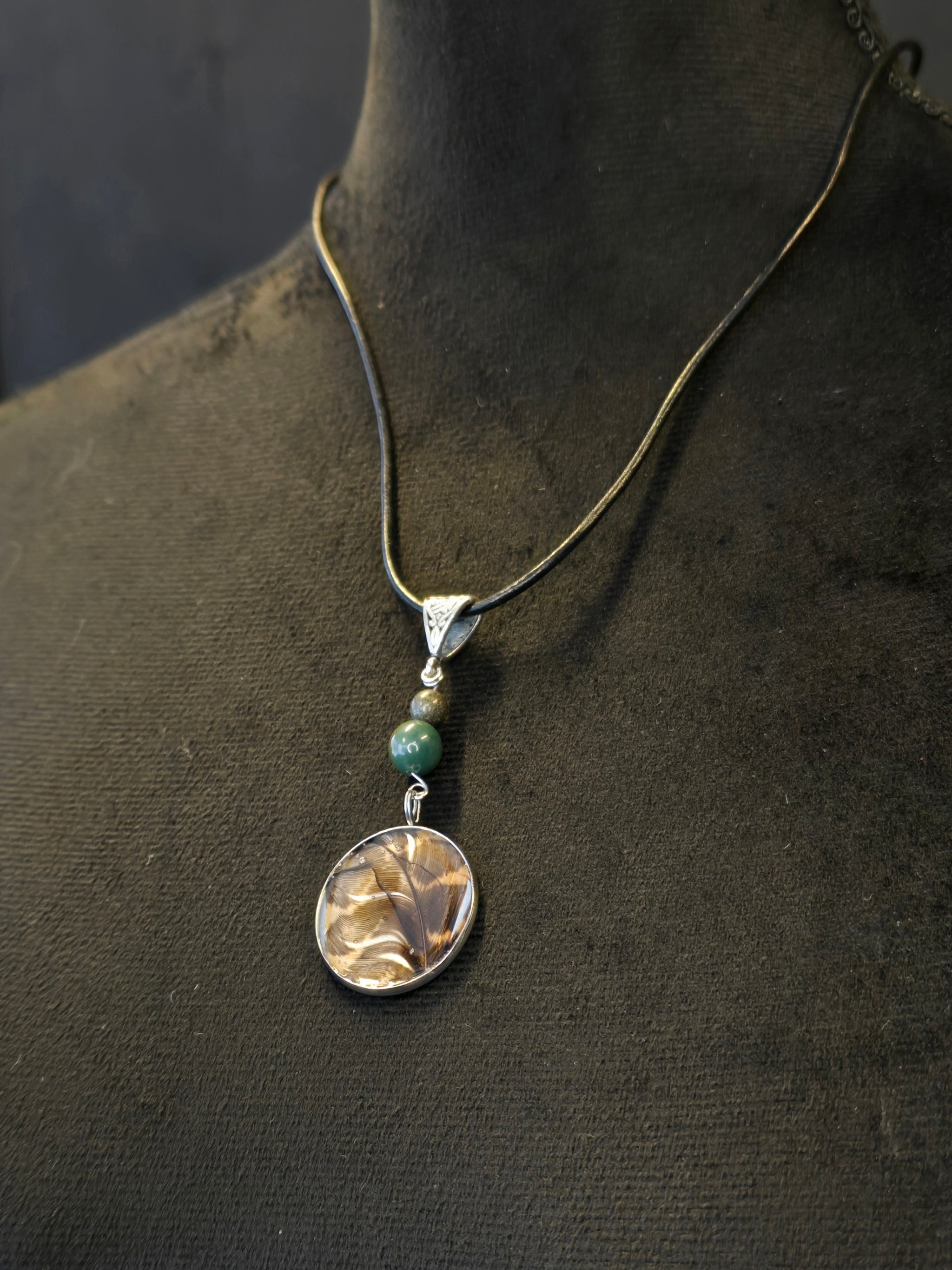 Metsän Kajo niminen amuletti, jossa on hartsi amuletin sisällä lehtokurpan sulka, ja sammalakaatti ja pyriittihelmet.